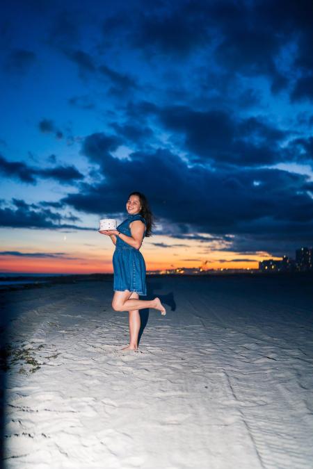 Woman balancing on one foot holding a birthday cake on the beach under a dramatic sky