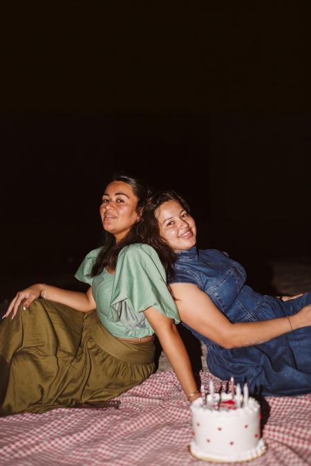 Two friends leaning together on a picnic blanket with a birthday cake on the beach at night