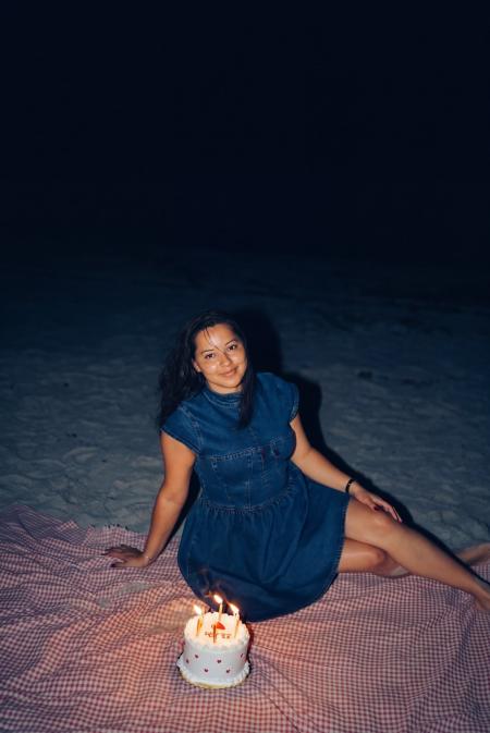 Woman in a denim dress on a pink picnic blanket beside a candle-lit birthday cake on the beach