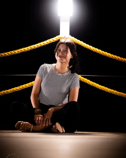 Model in a white tee sitting on a boxing ring canvas under a single spotlight