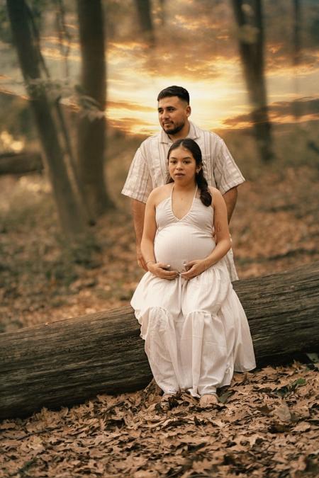 Expecting couple in an autumn forest at sunset, she sits on a fallen log and he stands behind her