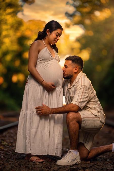 Father-to-be kneeling to kiss his partner on the belly on a railway track in warm golden light