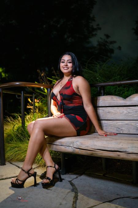 Woman in a red dress and heels seated on a wooden bench on a softly lit path at night