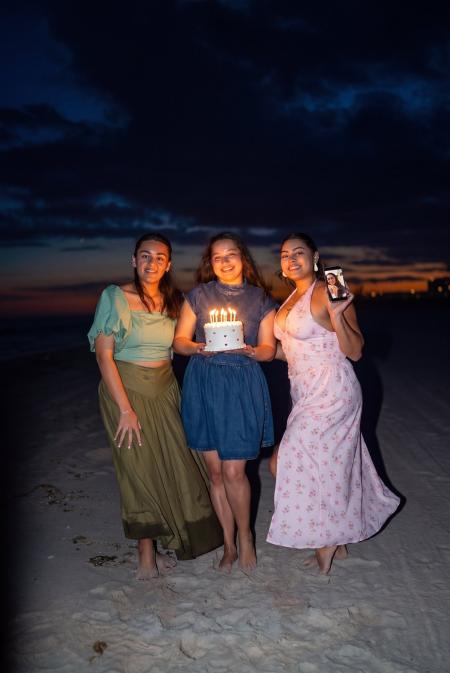 Three friends smiling with a birthday cake on the beach at dusk with an orange sunset behind them