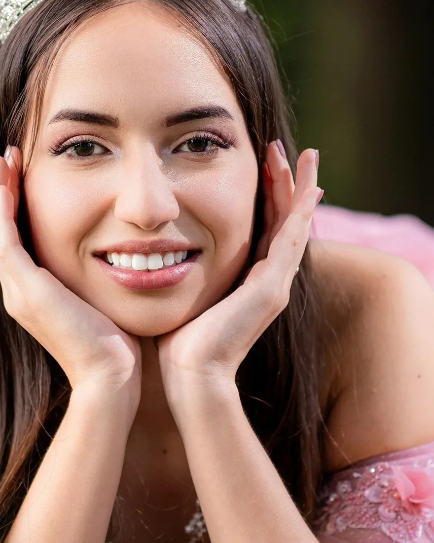 Smiling young woman in a tiara resting her face in her hands, a pink quinceañera gown around her