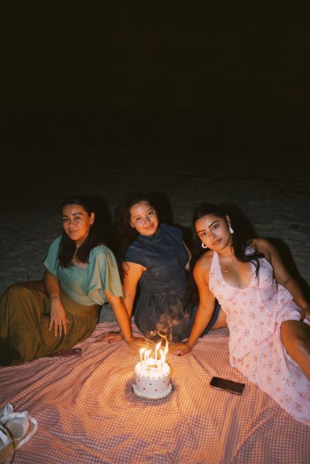 Three friends on a pink picnic blanket around a lit birthday cake on the beach at night