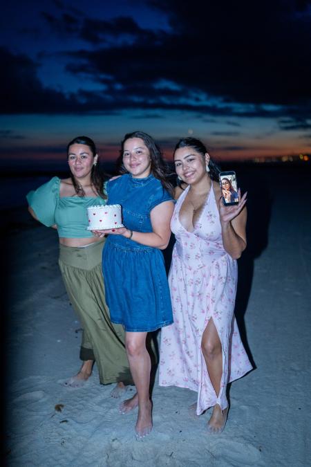 Three friends posing on the beach with a birthday cake and a sunset behind them