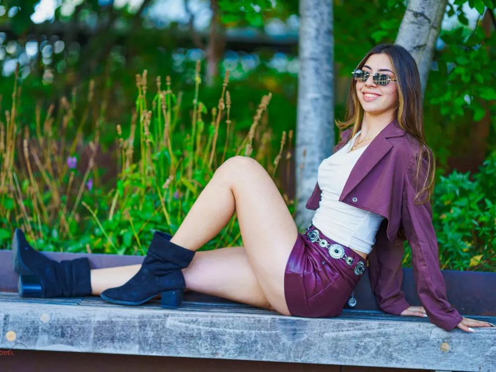 Woman in sunglasses and a burgundy jacket sitting on a park bench with plants and trees behind her
