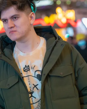 Young man in a green parka on a busy New York street at night, city lights behind him
